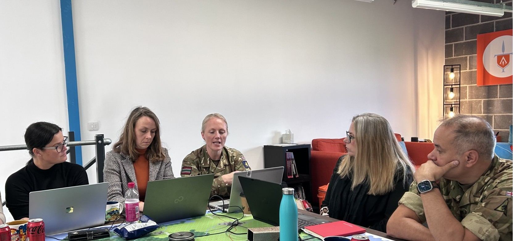 A mixed team of military officers and civilians confers over laptops and a hex map during a decision making exercise at Evocatus